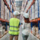 Work moment. Back view of elegant long-haired redhead woman and man in helmet and overalls standing communicating in aisle between warehouse shelves