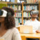 Diverse group of students watching virtual video tutorial in library. Man and women wearing virtual reality headsets, sitting at desks with laptops. Hardware for library concept